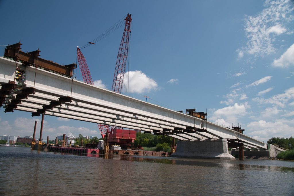 Large bridge under construction spanning across river with red cranes, steel beams, and industrial equipment visible on sunny day.