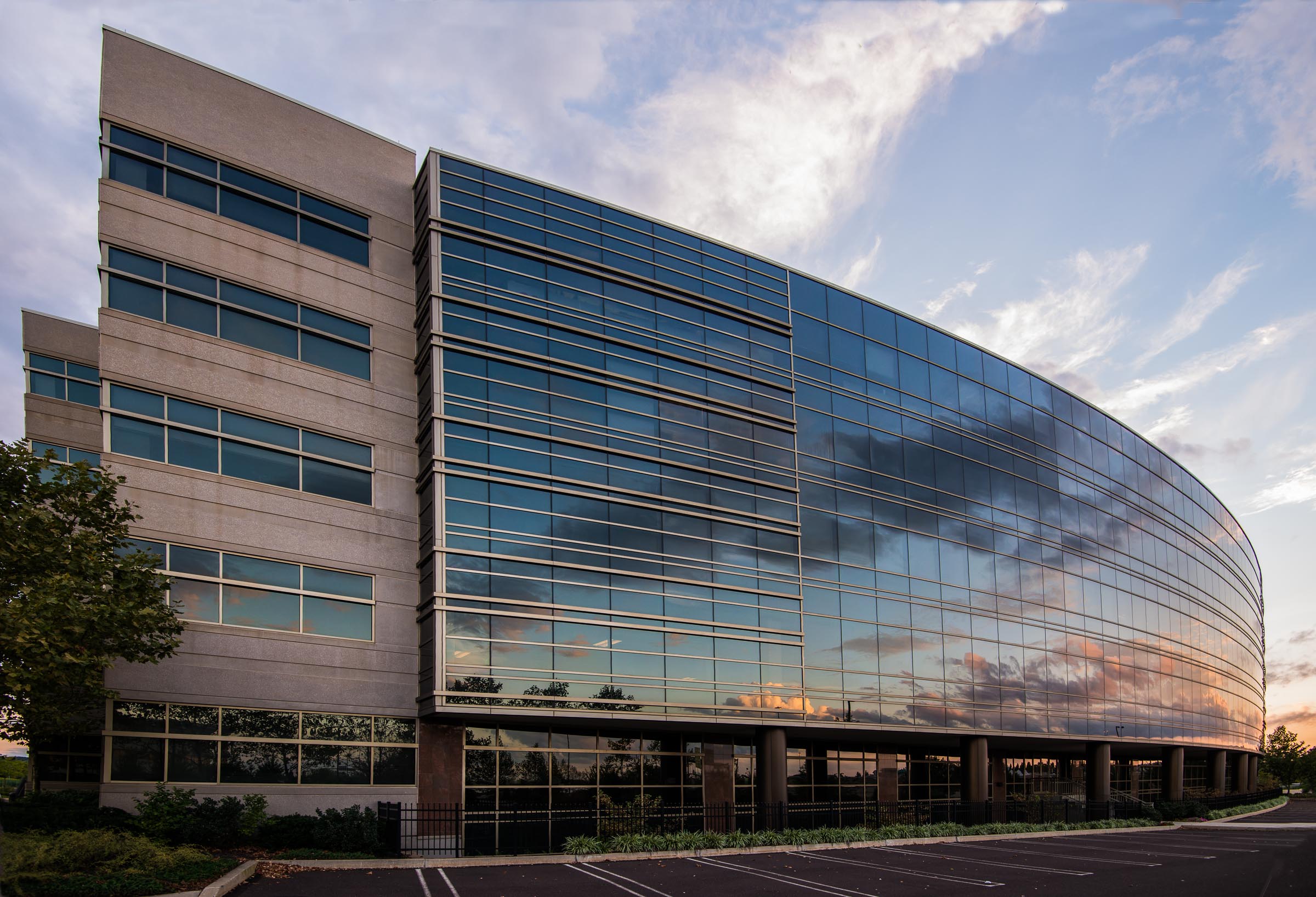 Modern curved glass and stone office building with horizontal window bands reflecting cloudy sky at sunset, featuring contemporary architecture design.