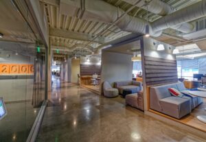 Modern office lobby with industrial ceiling, concrete floors, gray upholstered seating, and glass partitions showcasing contemporary workspace design.