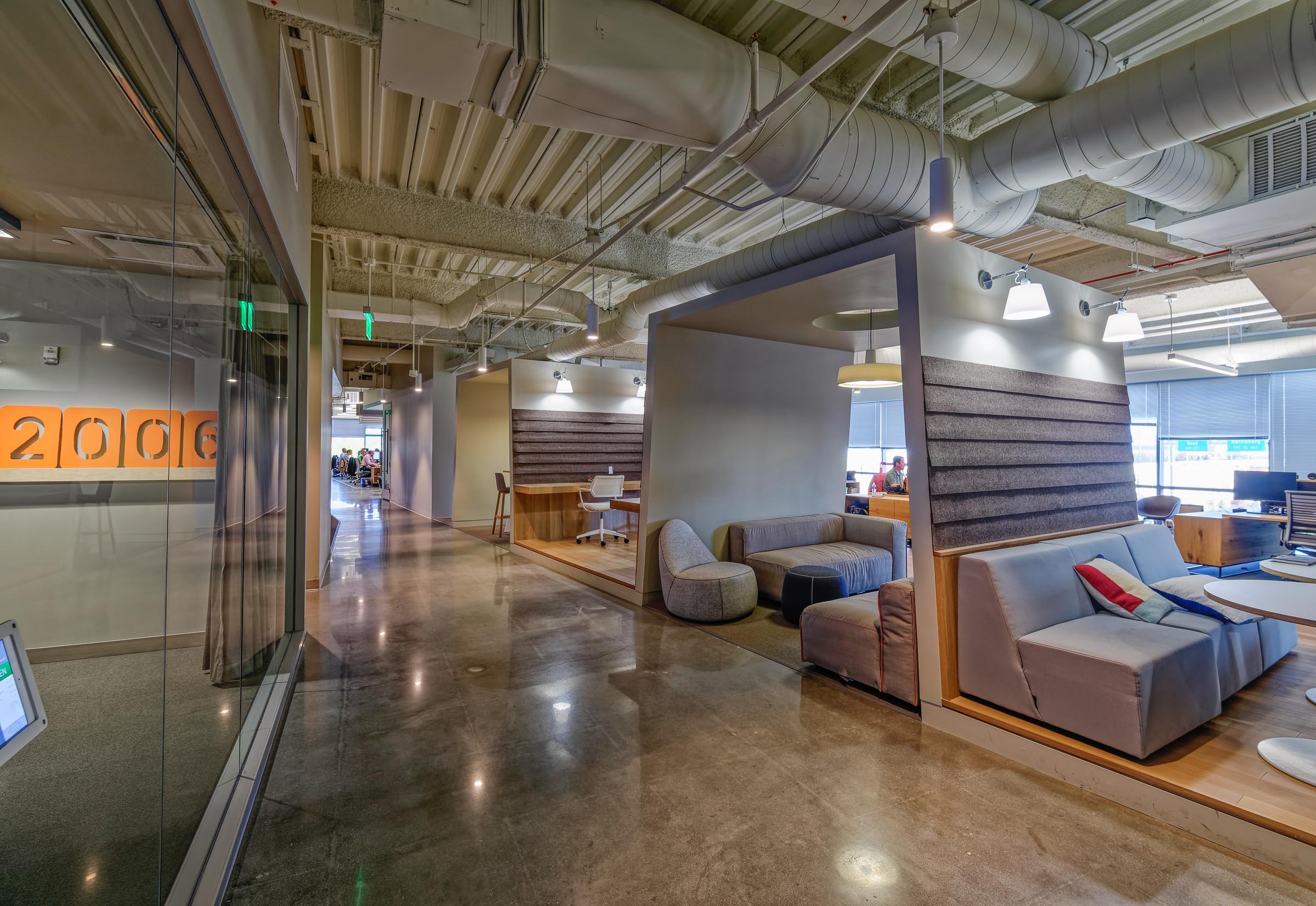 Modern office lobby with industrial ceiling, concrete floors, gray upholstered seating, and glass partitions showcasing contemporary workspace design.