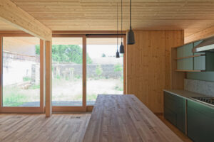 Modern wooden kitchen with large glass doors, dark pendant lights, green cabinetry, and stone courtyard view.