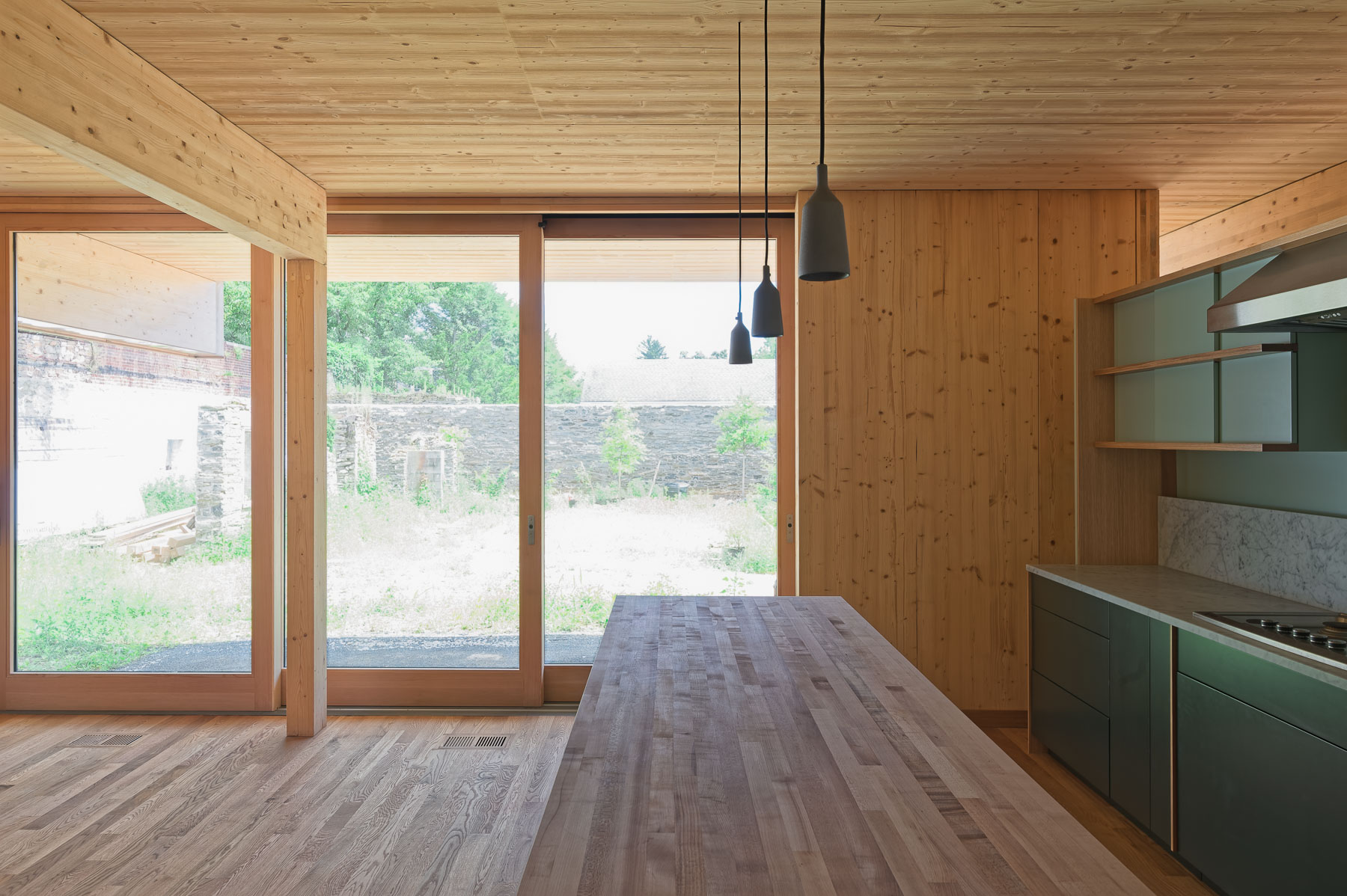 Modern wooden kitchen with large glass doors, dark pendant lights, green cabinetry, and stone courtyard view.