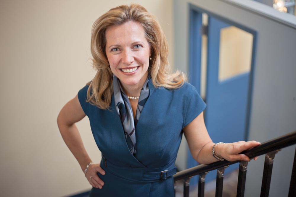 Professional woman in blue business attire smiling while holding stair railing in modern office hallway