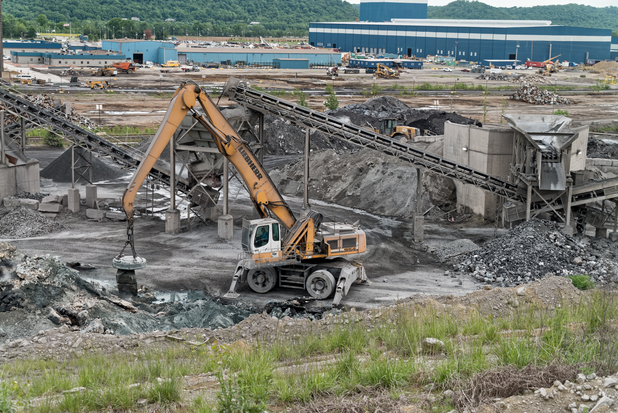 Yellow excavator with chain hook at active construction and demolition site with industrial buildings and processing equipment in background.