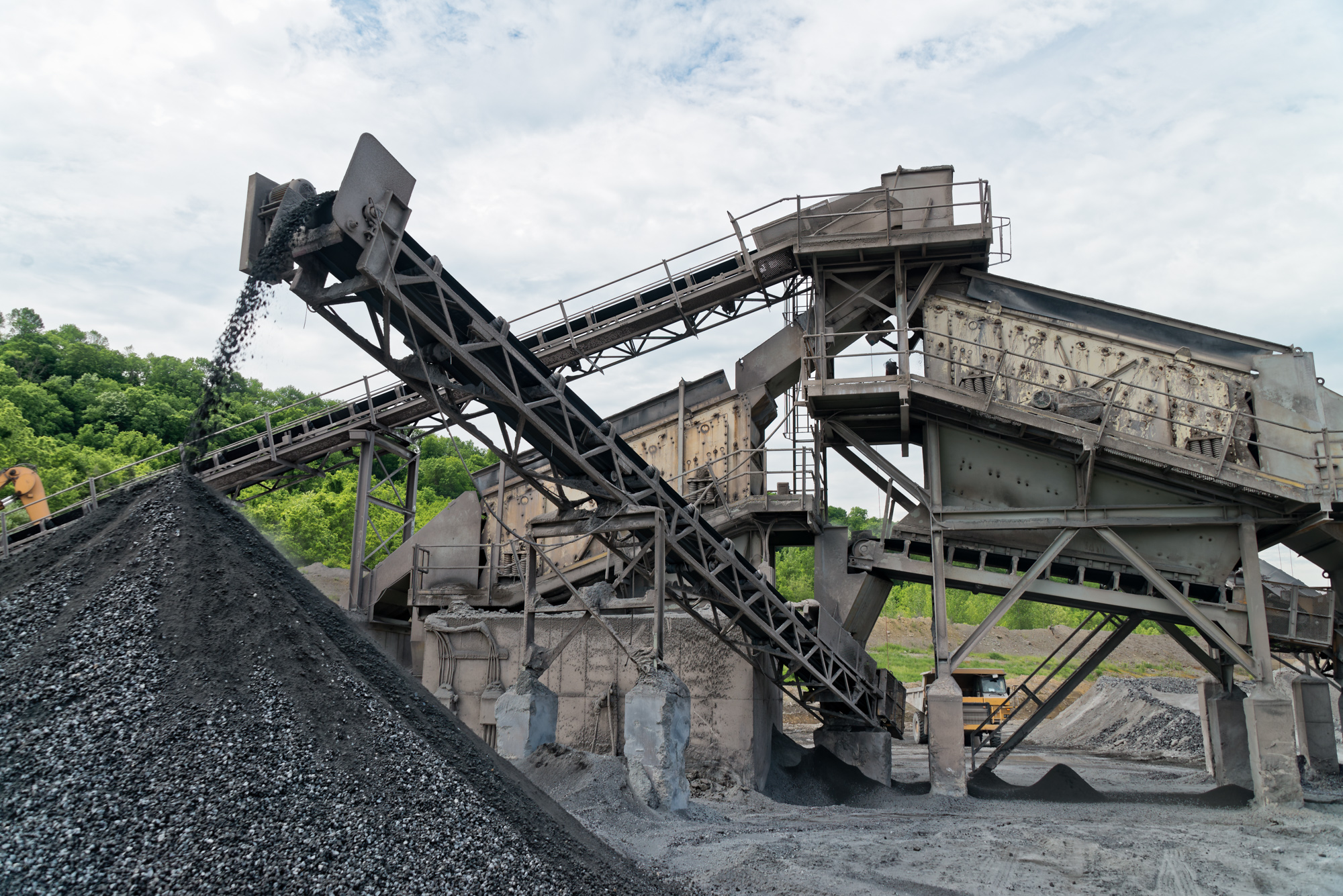 Industrial aggregate processing plant with conveyor belts, crushers, and stockpiles of crushed stone material against green hillside backdrop.