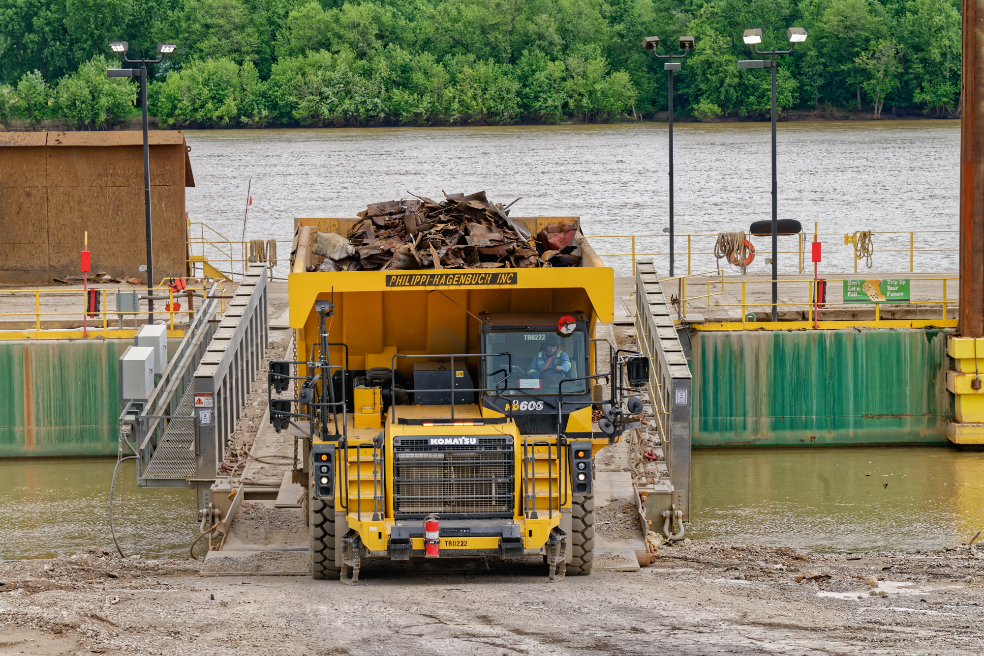 Yellow Komatsu dump truck loaded with scrap metal at industrial waterfront facility with river and green trees behind.