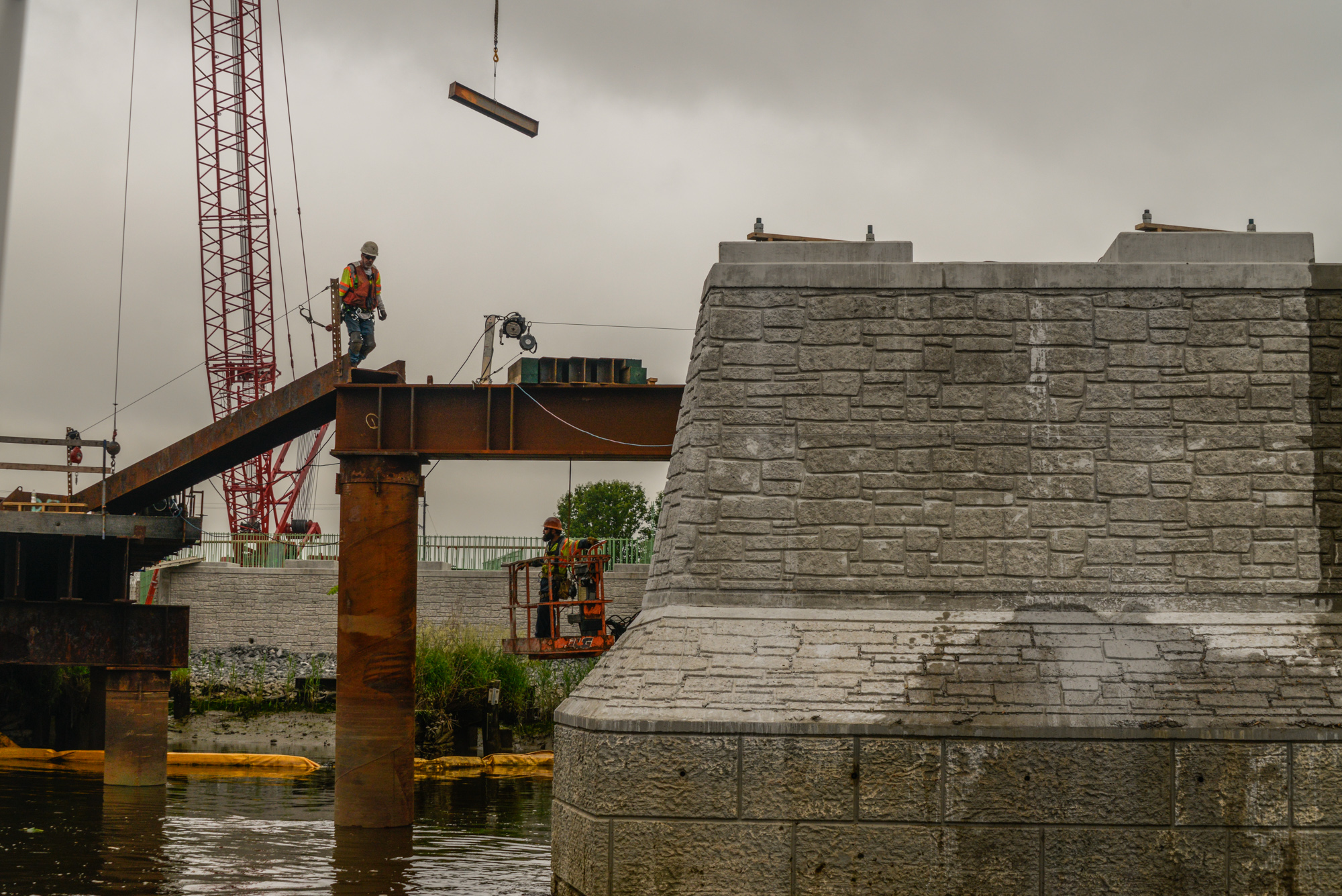 Construction workers operate crane and equipment at waterfront site with stone structure and red tower under overcast sky.