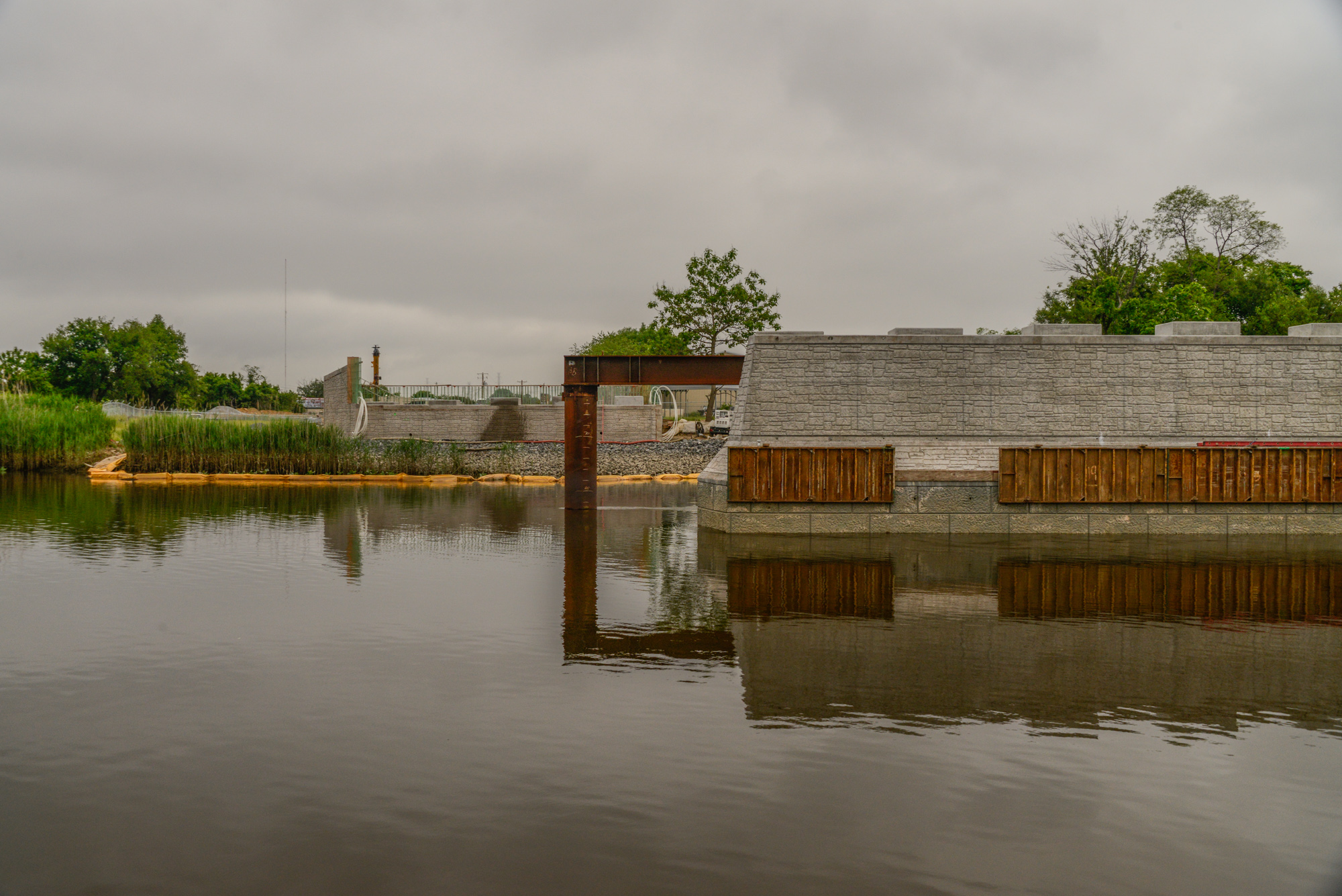 Modern industrial waterfront facility with concrete buildings, wooden dock structures, and calm water reflections under overcast sky.