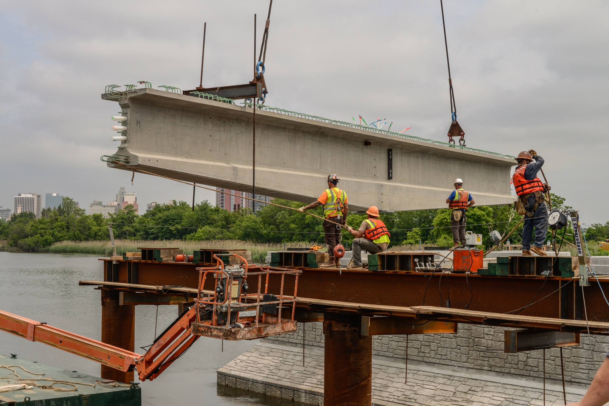 Construction workers in safety gear using cranes to lift a large concrete bridge beam over water at an industrial job site.