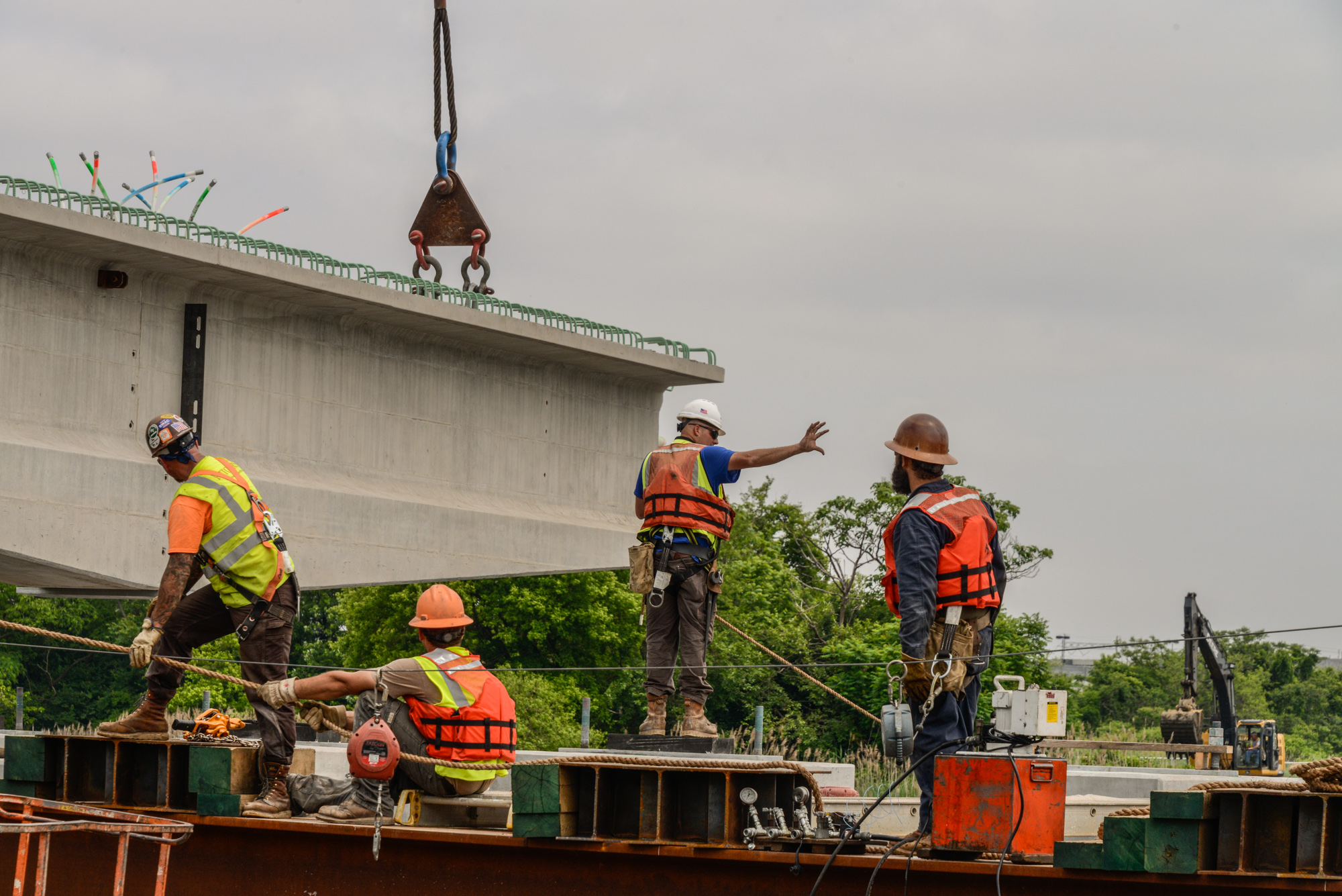 Construction workers in safety gear operating a crane to lift equipment on a barge beneath a concrete bridge structure.