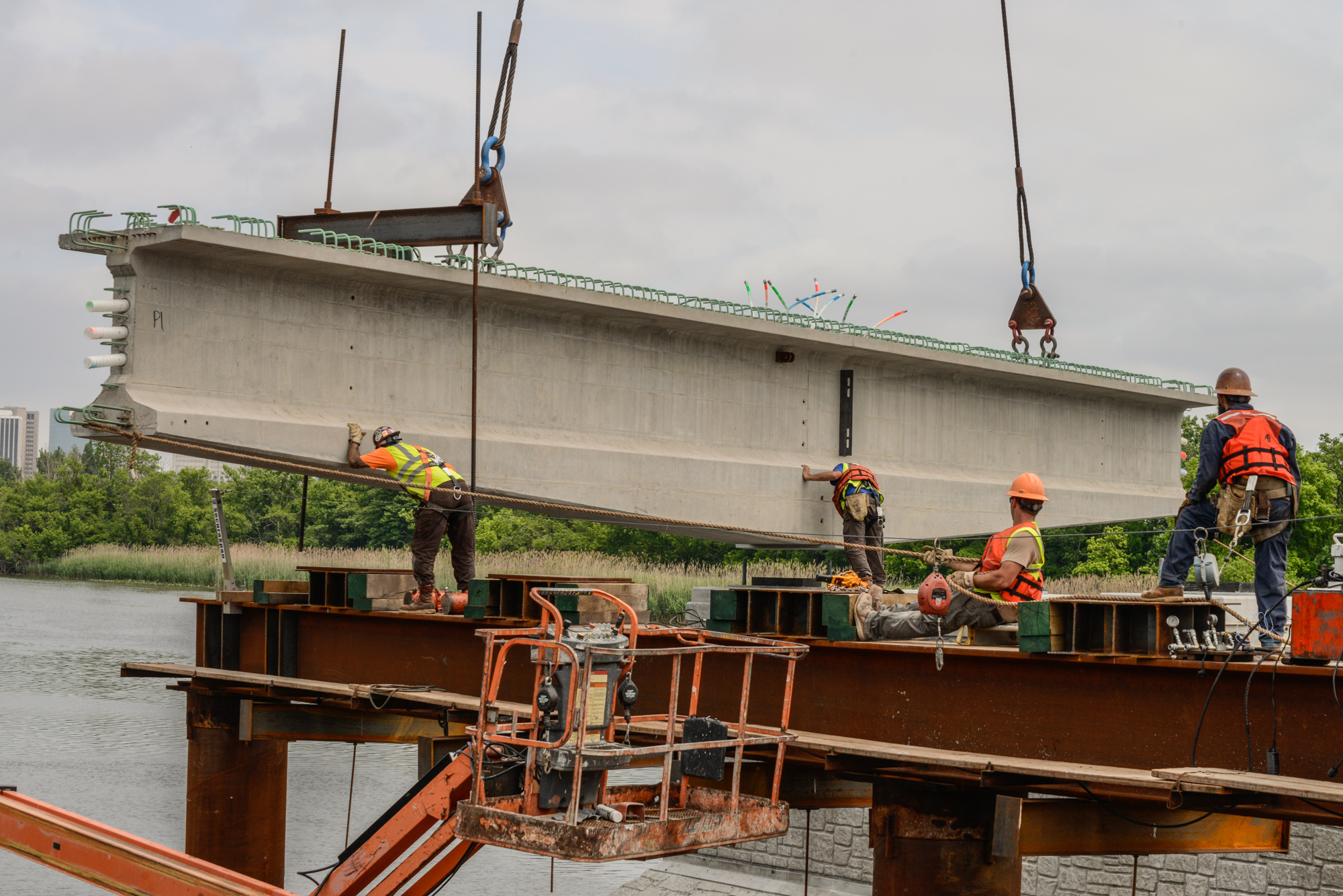 Construction workers in safety gear operating crane equipment to lift a large concrete bridge beam over water during industrial construction project.