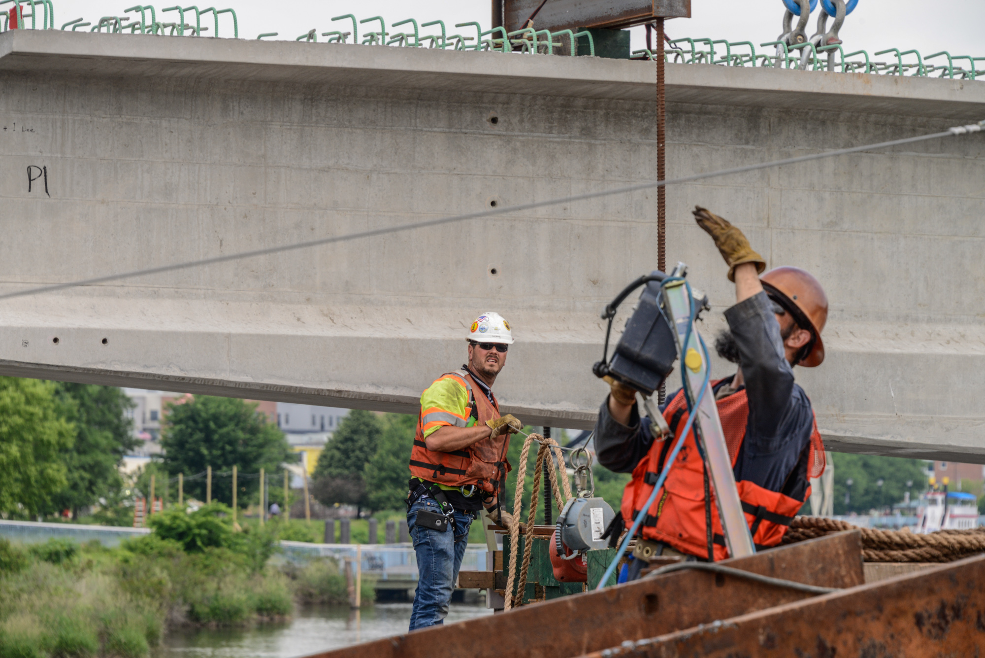 Construction workers in safety gear performing rope work on industrial bridge structure over water with rigging equipment.