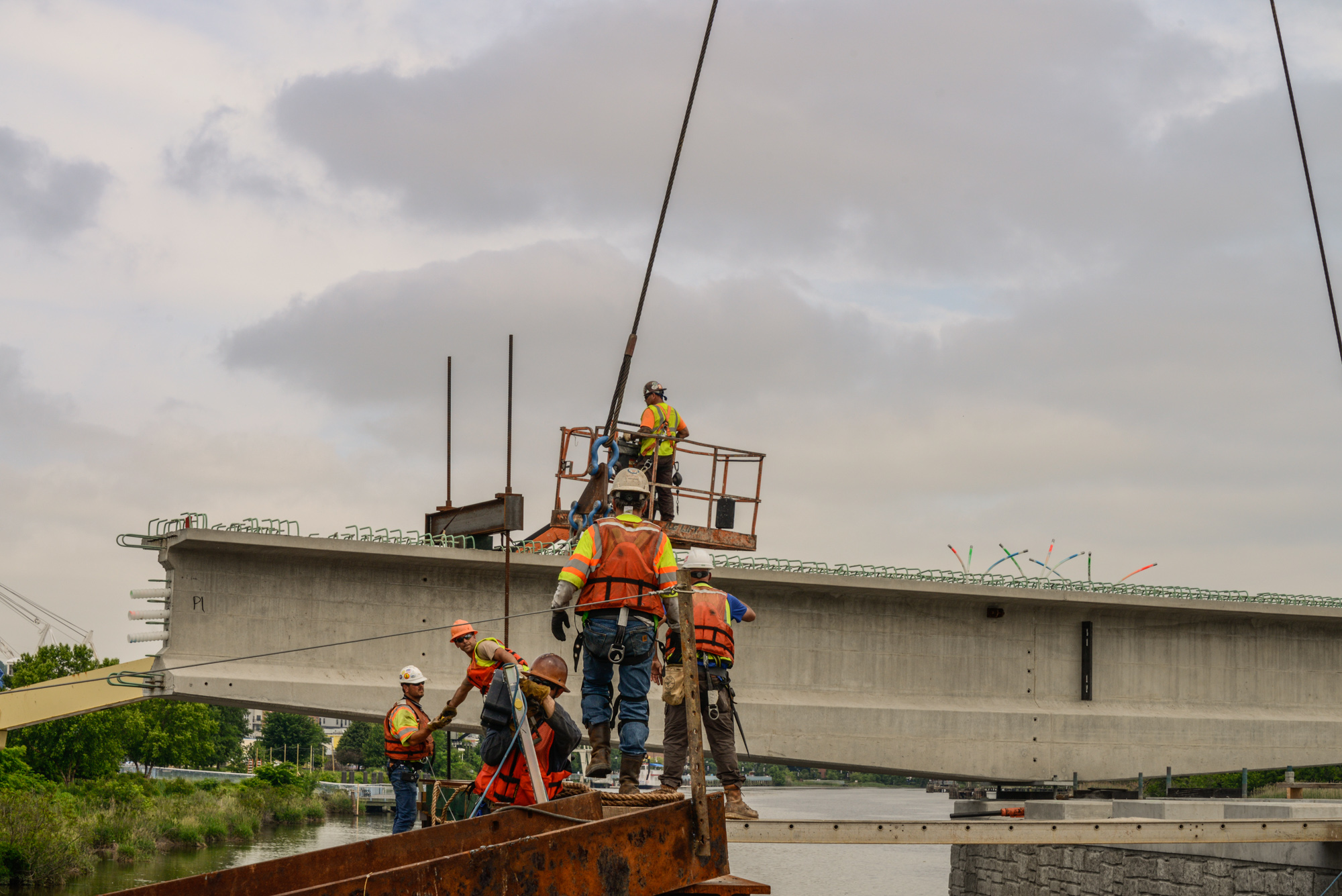 Construction workers in safety gear operating crane equipment on industrial bridge structure over water during infrastructure project.