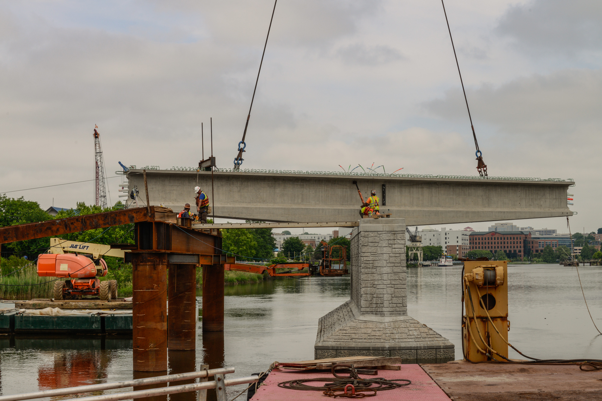 Large concrete bridge beam suspended by cables during industrial construction over river with workers and equipment.