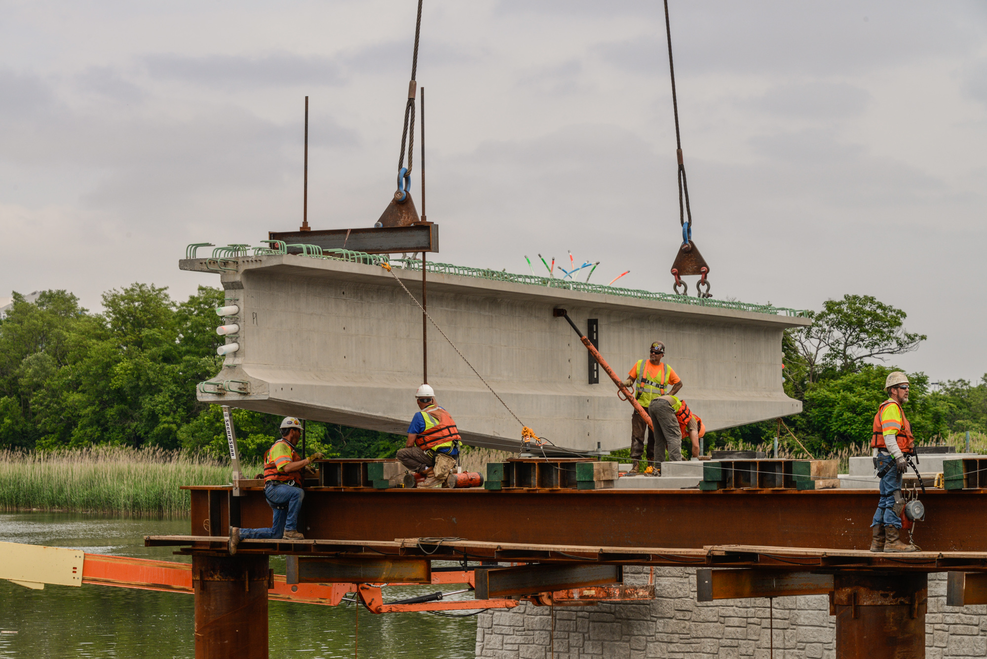 Construction workers in safety gear guide a large concrete bridge beam suspended by crane cables over water during industrial construction project.