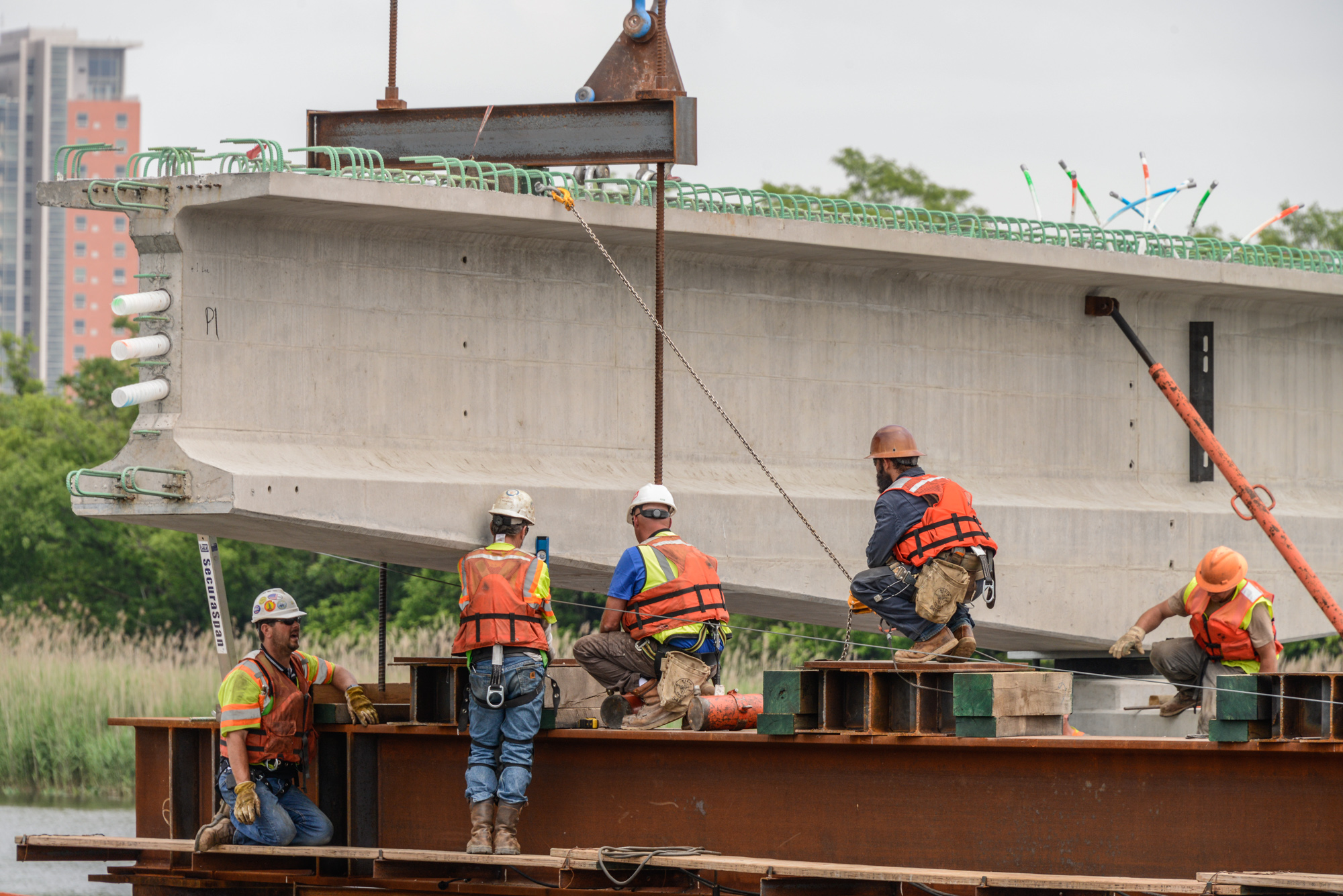 Construction workers in safety gear operating heavy machinery to lift a large concrete bridge beam at a waterfront industrial site.