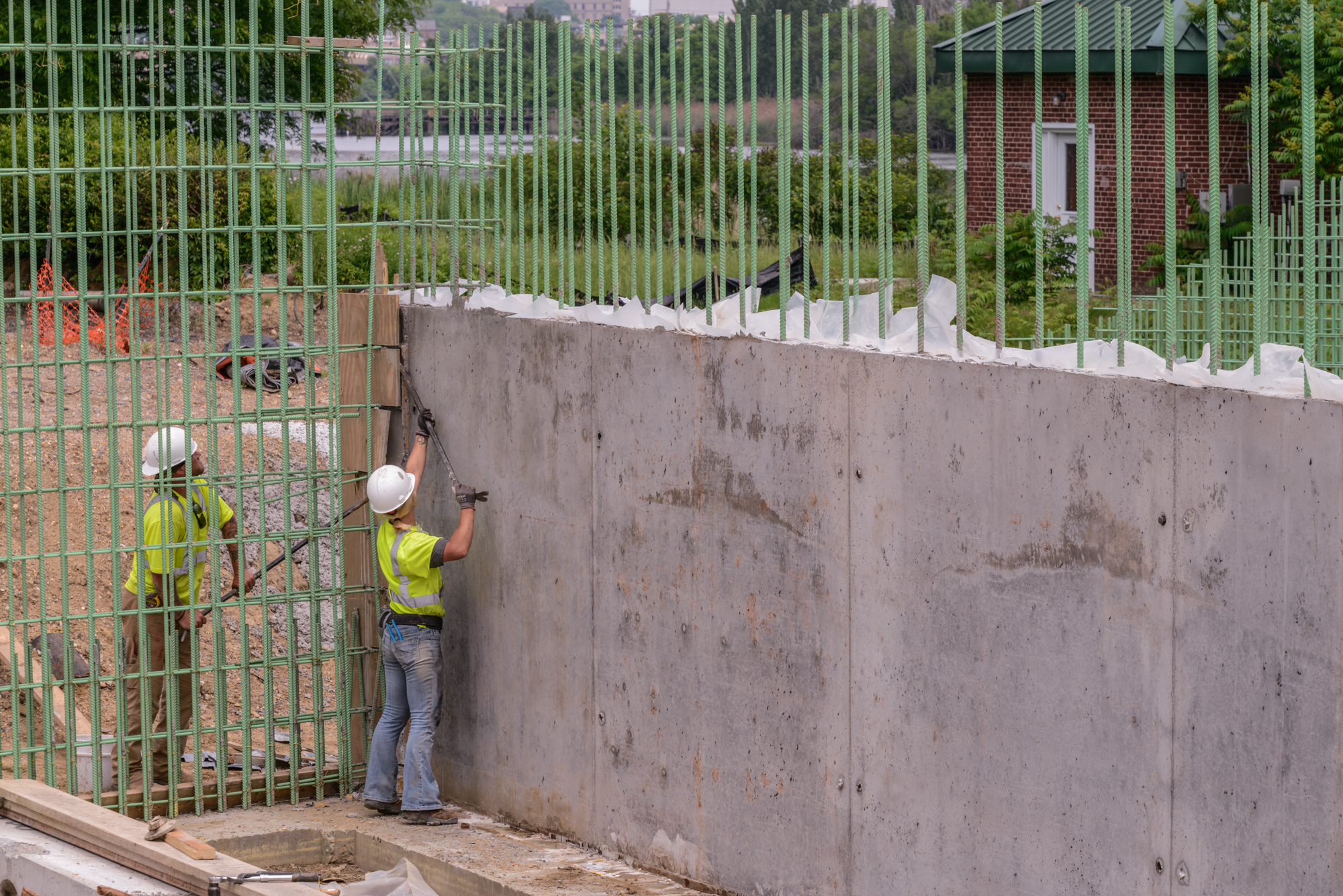 Construction worker in safety gear installing green metal fencing along concrete wall at industrial building site.
