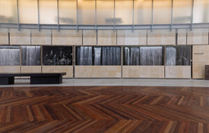 Modern gallery space with herringbone wood flooring, beige stone walls, and black and white art documentation photographs displayed on illuminated panels.