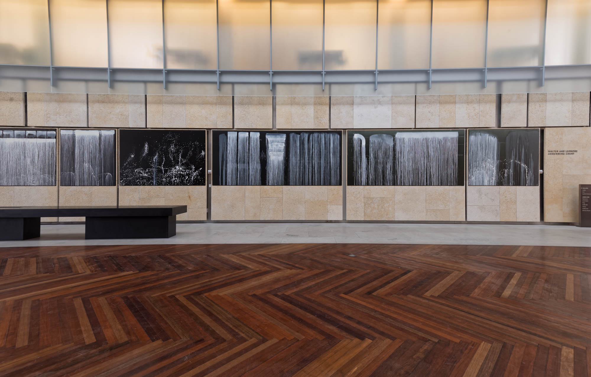 Modern gallery space with herringbone wood flooring, beige stone walls, and black and white art documentation photographs displayed on illuminated panels.