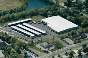 Aerial view of industrial warehouse complex with white roofs surrounded by green trees, river, and residential area