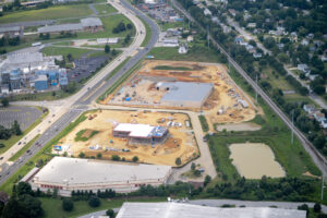 Aerial view of commercial construction site with multiple buildings under development, surrounded by highways, residential areas, and green spaces.