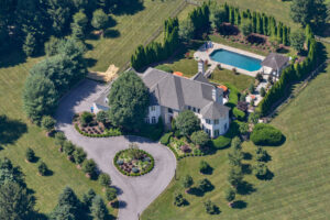 Aerial view of luxury estate with circular driveway, swimming pool, manicured grounds, and surrounding forest landscape.