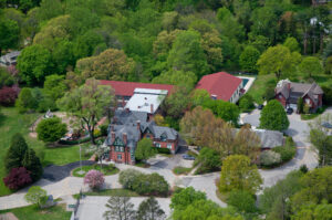 Aerial view of residential neighborhood with red brick Victorian mansion, modern homes with red roofs, lush green trees, and manicured lawns.