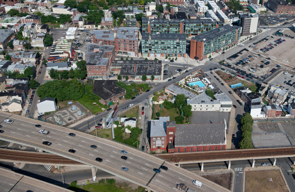 Aerial view of urban neighborhood with highway overpass, residential buildings, parking lots, and recreational facilities surrounded by green trees.