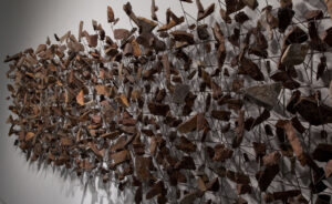 Dried brown leaves and seed pods suspended on thin stems against white wall, creating abstract botanical installation art sculpture.