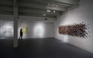 Contemporary art gallery exhibition with white walls displaying mixed media installations including colorful geometric patterns, linear drawings, and brown s…